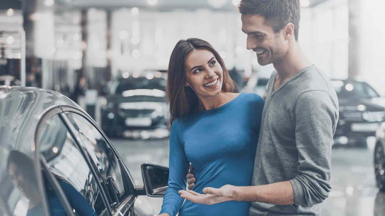A young couple standing in a car dealership, looking at a car and talking to a salesperson.